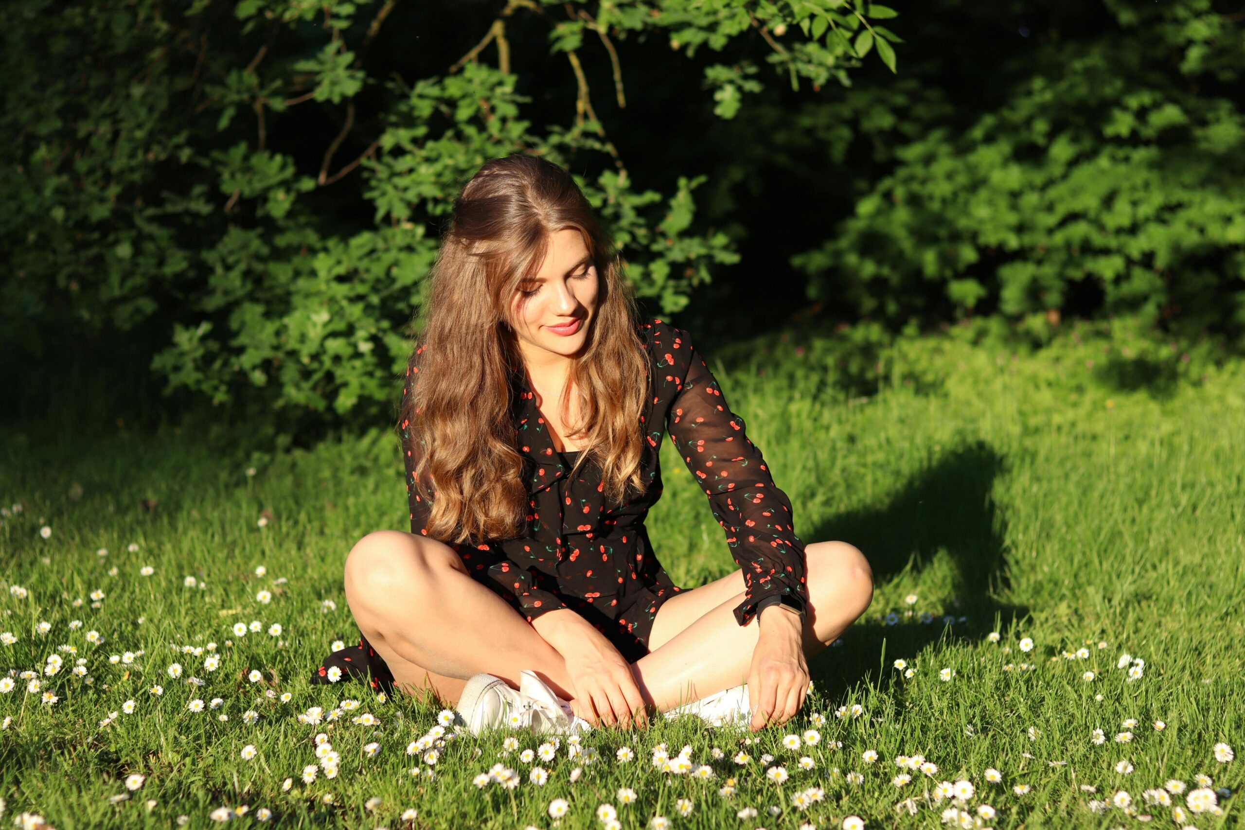 A woman in a black dress sits on grass in a sunlit field surrounded by daisies, exuding joy and relaxation.